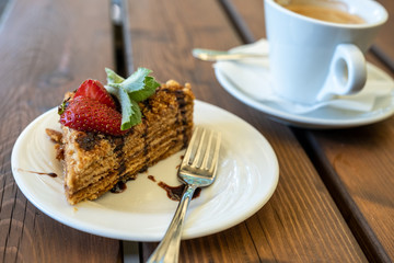 a cup of coffee and a piece of honey sponge cake decorated with strawberries and mint on a wooden table