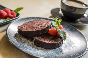 Chocolate swiss roll with jam decorated with raspberries on a plate and black coffee on the table. Selective focus.