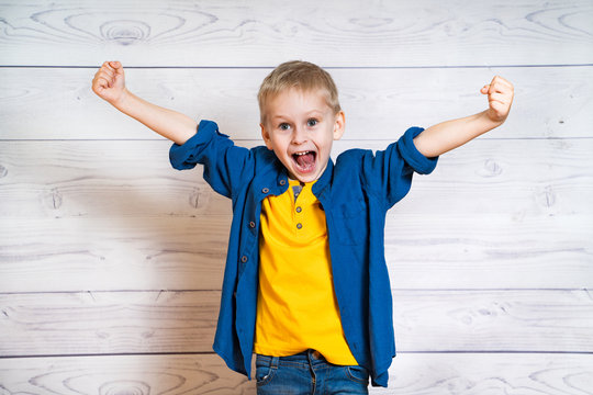 Little Boy In Yellow T-shirt And Blue Shirt Showing Happiness With Opened Mouth In Studio. Smiling Child With Spreading His Hands Isolated On The Light Wooden Background.