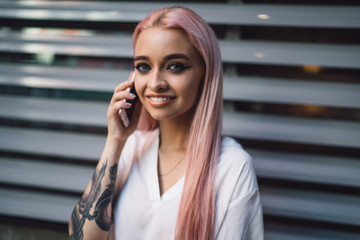 Close up portrait of positive female teenager with pink hair enjoying good internet connection on cellphone, happy woman with tattoo smiling at camera while phoning to best friend via smartphone app