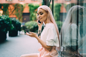 Positive female teenager dressed in casual wear taking rest on city street making international call to customer service, attractive millennial hipster girl with pink hair phoning via cellphone