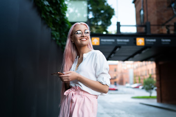 Cheerful woman in eyewear for vision correction smiling while waiting friend on urban area with mobile gadget in hands, happy millennial female blogger with pink hair excited with idea for publication