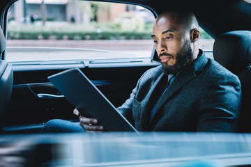 Businessman with black skin sitting on backseat in taxi automobile and searching contact number for making online video conference via modern touch pad, concept of market digitalisation on tablet