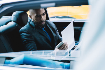 Dark skinned male strategist checking information for create productive plan for company development working on way to office building, business passenger in automobile reading contract details