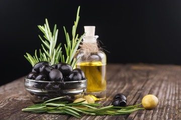 black olives, a small bottle of olive oil and a sprig of rosemary on a wooden table on a dark background
