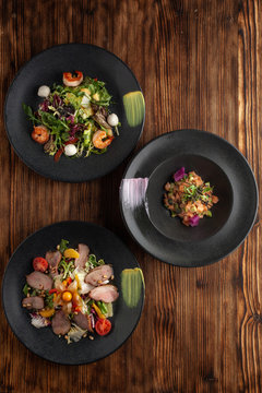 Variety Of Fancy Appetizers On A Minimalist Wooden Background: Raw Salmon Tartare With Avocado, Green Salad With Prawns, Duck Breast Salad With Orange Dressing