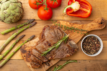 Marbled beef steak on a board with rosemary pepper, seasoning, and fresh vegetables, tomatoes and asparagus, artichoke, on a wooden background