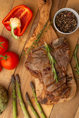 Marbled beef steak on a board with rosemary pepper, seasoning, and fresh vegetables, tomatoes and asparagus, artichoke,