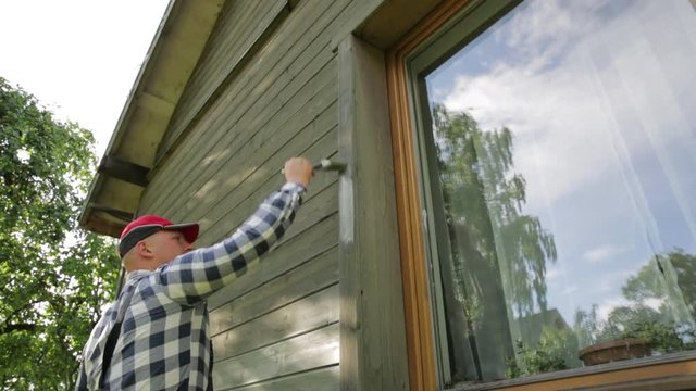 Man worker painting wooden house exterior wall with paintbrush and wood protective color