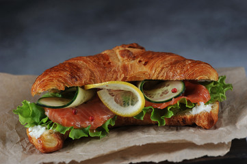 Croissant as a sandwich. Soft cheese, weak salt salmon, lettuce, cucumber and lemon slices are included in the filling. Dark background and craft paper. Close-up.