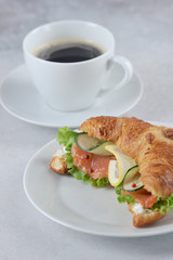 Croissant as a salmon sandwich and a cup of coffee. Soft cheese, salted salmon, salad, cucumber and lemon slices are included in the sandwich filling. Light background. Close-up. Vertical orientation.