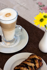 Tomorrow. Cappuccino in a tall glass with white foam and cinnamon, butter roll with chocolate and yellow chrysanthemums in a white vase.