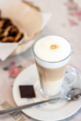 Cappuccino in a tall glass with a high white foam with cinnamon. Next to the plate is a long teaspoon and a square slice of chocolate. In the background in the basket is a fresh bun with chocolate.