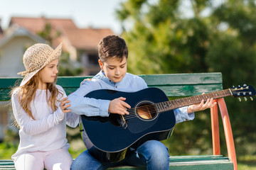 Young girl and boy with the guitar sitting at the bench in park. Children love. 