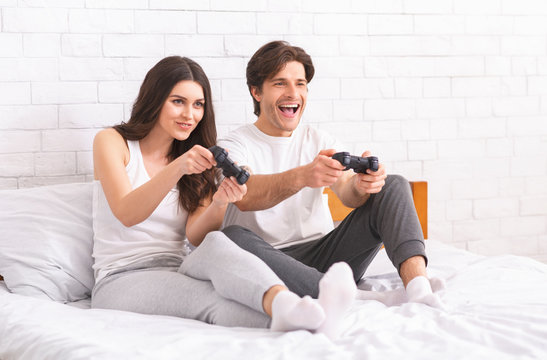 Emotional millennials playing video game, sitting with joystick on bed