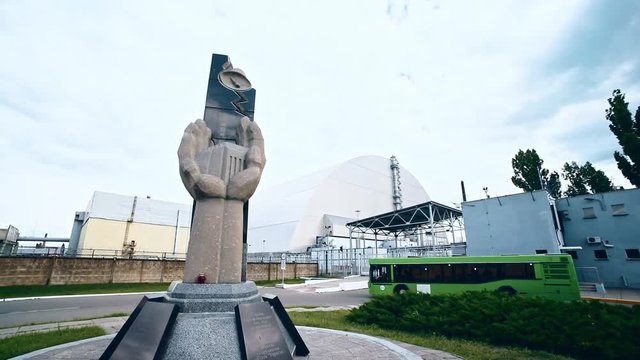Wide-angle view of Chernobyl plant courtyard with memorial statue to nuclear disaster explosion liquidators in summer day. Travel bus with tourists arriving at the power station.