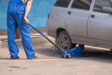 Man in blue workwear jacks up a car. Wheel repair. Metal key. The concept of road workshop, minor...