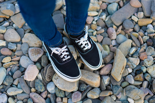 Women's legs in jeens and black sneakers on pebbles.