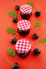 Cherry jam. Homemade sweet cherry jam in  glass jars set row and ripe sweet cherry berries against a bright red background. View from above.