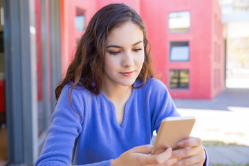 Concentrated student girl texting message on smartphone. Young woman in blue sweater using gadget outdoors. Online communication concept