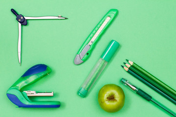 Color pencils, pen, apple, felt-tip pen, paper knife, compass and stapler on green background.