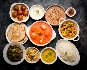 Assorted Indian food like paneer butter masala, dal, roti, rice, sabji, gulab jamun and bound raita served in bowls over moody background, selective focus