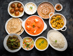 Assorted Indian food like paneer butter masala, dal, roti, rice, sabji, gulab jamun and bound raita served in bowls over moody background, selective focus