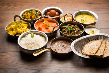 Assorted Indian food like paneer butter masala, dal, roti, rice, sabji, gulab jamun and bound raita served in bowls over moody background, selective focus