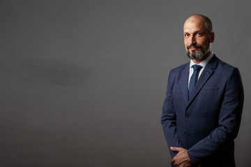Handsome young bearded businessman in classic suit is adjusting his jacket and looking away, on a gray background