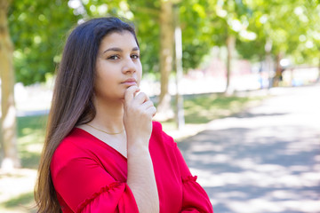 Dreamy pretty young woman touching chin in park. Beautiful lady wearing red blouse and looking at camera with green trees in background. Contemplation and nature concept.