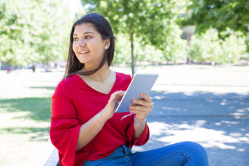 Happy young woman sitting and using tablet in park. Beautiful lady wearing red blouse with green trees in background. Technology and nature concept.