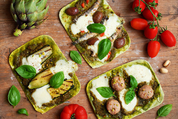 Top view of three small green pizza with pesto sauce, grilled zucchini, sardines and meatballs, mozzarella cheese and basil on wooden table. Italian snack.