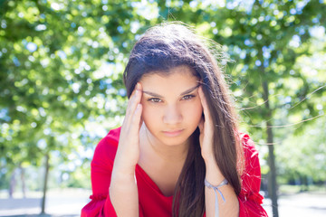 Serious young woman touching temples and concentrating in park. Beautiful lady wearing red blouse and looking at camera with green trees in background. Contemplation and nature concept. Front view.