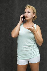 portrait above the knee of a beautiful blonde woman girl with short curly hair on a gray background in bright linen talking on the phone and showing a lot of emotions. Experienced model shows hands.