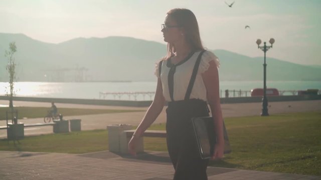 Caucasian young business woman in a white shirt and glasses goes, holding in hand a laptop, backdrop the mountains and the sea. The average plan. The effect of illumination lit sun.