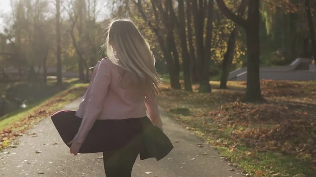 Beautiful girl in pink jacket turns around and smiles looking at camera outdoor in slow motion. Portrait of blonde woman spinning at sunset. Sun is shining. Medium shot