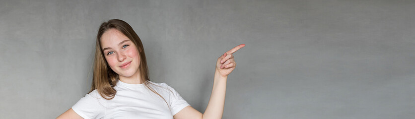  Portrait with copy space fashionable girl of the millennial generation z in white t-shirt pointing with index finger at empty space, looking at camera, isolated on gray background