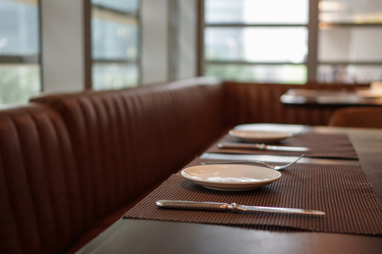 Plates, Forks And Knifes On Brown Table Mat And Leather Seat Near Window In Daylight