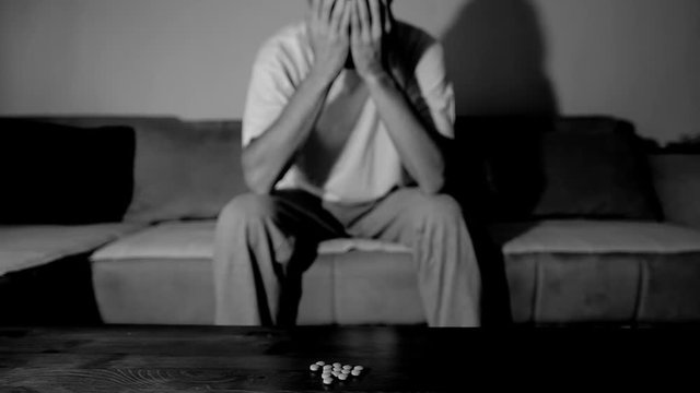 Depressed man suffering from suicidal depression want to commit suicide by overdose taking strong medicament drugs and pills pain killers sitting in his dark room selective focus moody dramatic look