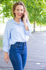 Joyful beautiful girl walking in park and talking on phone. Happy young woman in casual standing on walkway in park, calling on cell and smiling at camera. Telecommunication concept