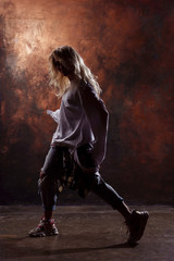 Fototapeta na wymiar Side photo of slim blonde dancer in torn jeans and sneakers on dark brown background