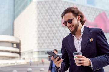 Photo of smiling man in sunglasses with phone in his hand while walking through city