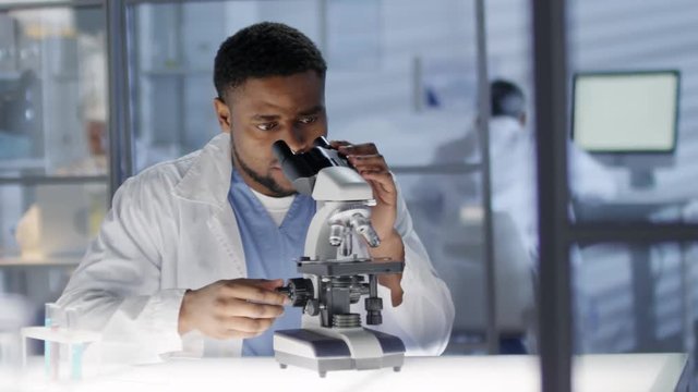 Waist-up shot of 30-something Afro-American technician, dressed in scrubs and white coat, sitting in hospital laboratory, studying tissue samples under microscope and turning adjustment knob