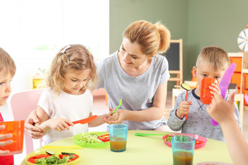 Nursery teacher with cute little children during lunch in kindergarten