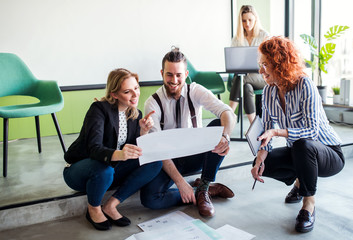 A group of young business people sitting on the floor in an office, talking.