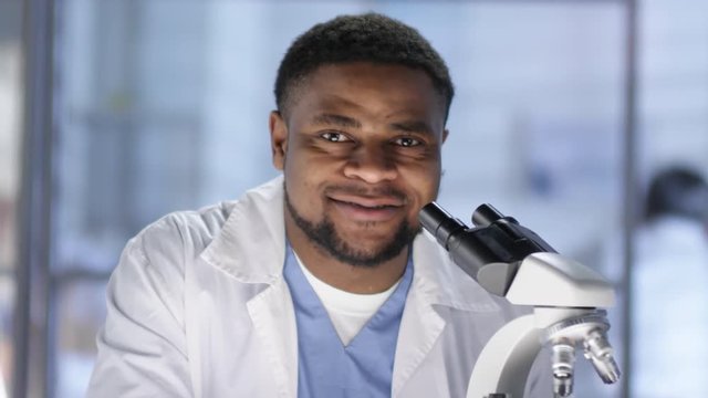 Chest-up portrait shot of middle-aged black lab technician, dressed in white coat, peering into microscope and studying tissue sample, then looking at camera with happy and delighted smile