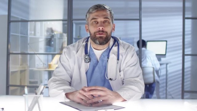 POV shot of middle-aged Caucasian male GP, dressed in scrubs and lab coat with stethoscope, sitting at light table in surgery and seeing patient, asking for medical history, diseases and complaints