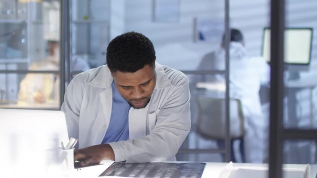 Waist-up shot of concerned Afro-American neurologist, dressed in lab coat and medical uniform, hurriedly studying MRI scan and typing on laptop for patient case record