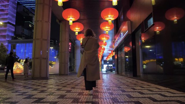 Caucasian girl in a long light raincoat, stands in the middle of the night on Macao street. Nearby are people. Above the girl hang red lanterns with yellow fringe.