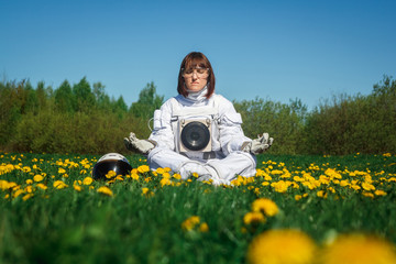 Beautiful girl astronaut without a helmet sits on a green lawn among flowers in a meditative...